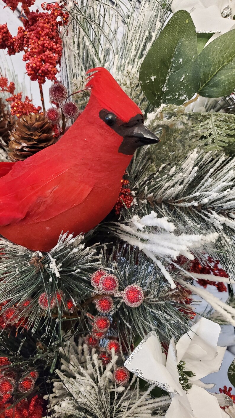 May include: A vibrant red cardinal bird ornament perched amidst frosted evergreen branches, red berries, and pinecones. The bird has a black beak and eye, and is surrounded by white poinsettia flowers. A festive holiday decoration.