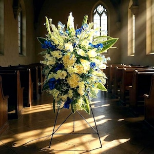 May include: A large floral arrangement on a black metal stand. The arrangement features white, yellow, and blue flowers with green foliage. The background is a church interior with pews and sunlight streaming through the windows.