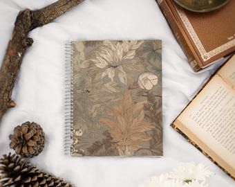 May include: A spiral bound notebook with a brown and green floral pattern. The notebook is lying on a white surface with a few pine cones and white flowers.