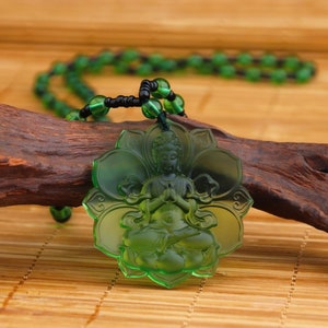 Peut inclure: Un collier avec un pendentif en verre vert représentant une sculpture détaillée d'un Bouddha assis en position de lotus. Le collier est composé de perles de verre vert et d'un cordon noir.