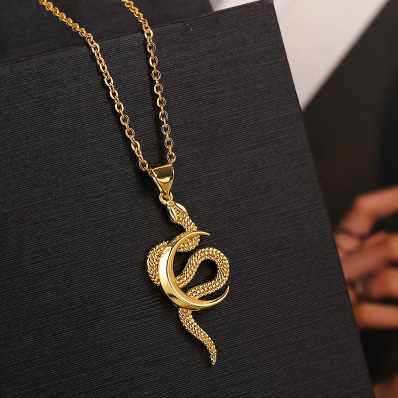 May include: A gold chain necklace with a pendant featuring a snake wrapped around a crescent moon.