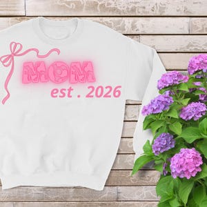 Peut inclure: Sweat-shirt blanc avec le mot "MOM" en lettres roses néon, un nœud rose et le texte "est. 2026". L'image présente également un buisson d'hortensias violets.