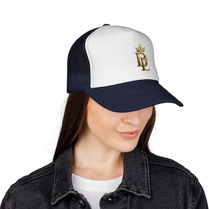 Peut inclure: Casquette de camionneur bleu marine et blanche avec une couronne dorée et les lettres "DL" en or. Le chapeau a un dos en filet et une visière incurvée. La personne porte une veste en jean noire.