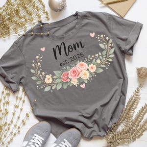 Peut inclure: T-shirt gris avec le mot "Mom" et "est. 2026" en noir, entouré d'une couronne florale de roses roses et crème, de feuilles vertes et de petits cœurs roses. Des baskets grises sont visibles en bas du t-shirt plié.