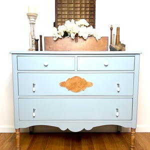 May include: A light blue dresser with four drawers and a decorative wooden accent. The dresser has white knobs and pulls, and sits on wooden legs with wheels. A wooden box with white flowers and other decorative items are on top.