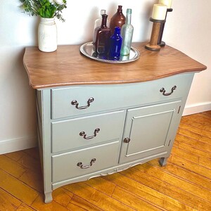May include: A light blue painted cabinet with a wooden top, featuring three drawers and a cabinet door. The cabinet is adorned with antique-style hardware and holds decorative bottles on a silver tray. A mason jar with white flowers is on the left.
