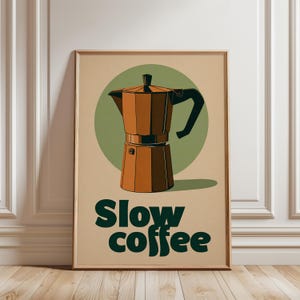 May include: A framed wall art print featuring a brown moka pot centered over a sage green circle on a beige background. Below the illustration, the text reads "Slow coffee" in bold, dark green, rounded, lowercase-style typography.