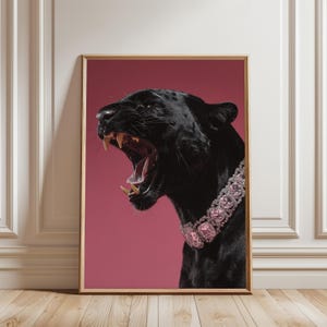 May include: A framed art print featuring a black panther with an open mouth, revealing its teeth. The panther wears a pink jeweled collar against a pink background. The artwork is displayed in a room with a wooden floor and white wall.