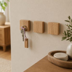 May include: Three square light brown wooden wall hooks mounted horizontally. The leftmost hook holds a silver metal key ring with a small wooden tag attached by a thin purple cord. The wood grain is visible on each of the three square blocks.