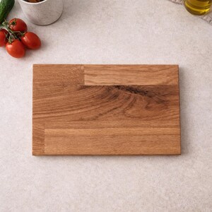 Pu&ograve; includere: Un tagliere rettangolare in legno con un motivo a venature naturali. La tavola &egrave; circondata da utensili da cucina, pomodori, un cetriolo, olio d'oliva e saliere e pepiere. La tavola misura circa 30 cm di lunghezza e 20 cm di larghezza.