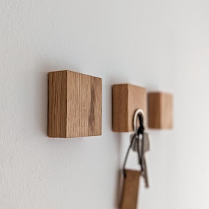 Drei Schlüsselhalter aus Holz an einer weißen Wand. Die Schlüsselhalter sind quadratisch und aus hellem Holz gefertigt. Ein Schlüsselhalter hat einen Satz Schlüssel daran hängen. Die Holzmaserung ist auf den Schlüsselhaltern sichtbar.