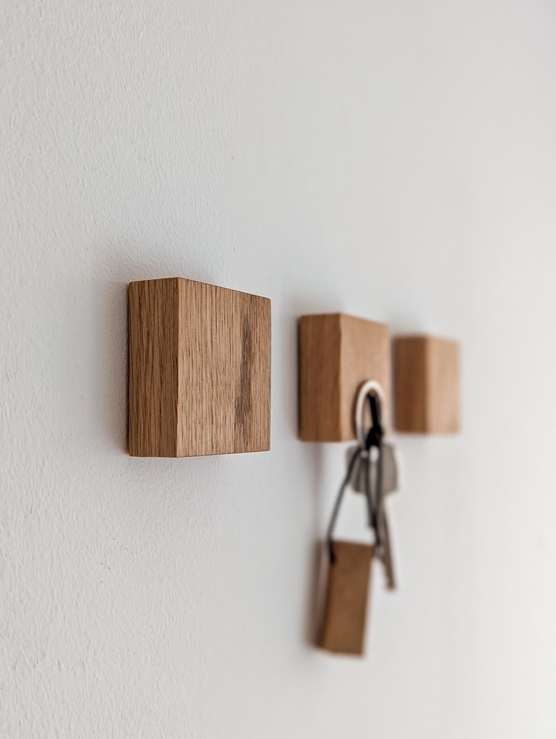 Drei Schlüsselhalter aus Holz an einer weißen Wand. Die Schlüsselhalter sind quadratisch und aus hellem Holz gefertigt. Ein Schlüsselhalter hat einen Satz Schlüssel daran hängen. Die Holzmaserung ist auf den Schlüsselhaltern sichtbar.