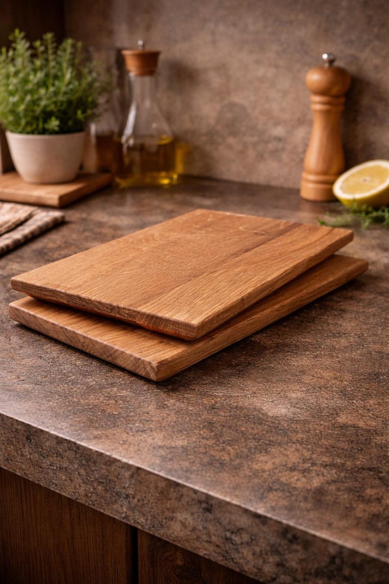 Pu&ograve; includere: Due taglieri rettangolari in legno impilati su un piano di lavoro marrone. Le tavole hanno una venatura naturale del legno e un colore marrone chiaro. L'ambiente della cucina comprende una pianta, una bottiglia d'olio e un limone.