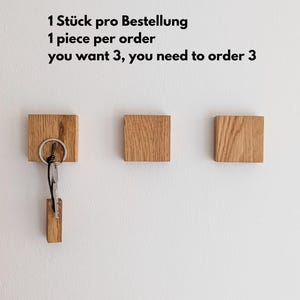 Drei hölzerne Schlüsselhalter an einer weißen Wand. Ein Schlüsselhalter hat Schlüssel befestigt. Der Text an der Wand lautet: "1 Stück pro Bestellung, wenn Sie 3 wollen, müssen Sie 3 bestellen."