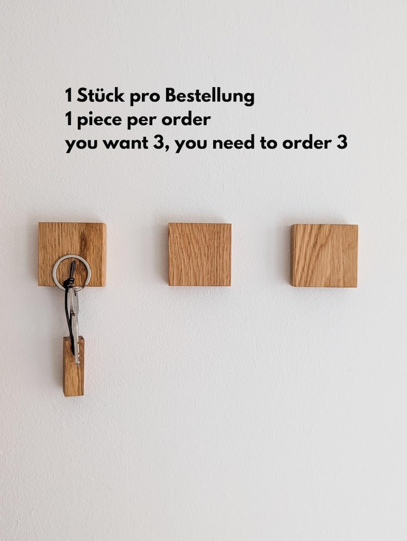 Drei hölzerne Schlüsselhalter an einer weißen Wand. Ein Schlüsselhalter hat Schlüssel befestigt. Der Text an der Wand lautet: "1 Stück pro Bestellung, wenn Sie 3 wollen, müssen Sie 3 bestellen."