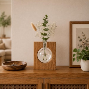 Könnte beinhalten: Eine klare Glaskolbenvase in einem rechteckigen Holzrahmen mit kreisförmigem Ausschnitt. Die Vase enthält einen Eukalyptuszweig, Schleierkraut und einen einzelnen beigen Trockengraswedel. Das Holz hat eine sichtbare Maserung und einen warmen Ton.