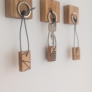Può includere: Tre portachiavi in legno montati su una parete bianca. Ogni supporto è un blocco quadrato di legno con un anello di metallo e un cordino nero. Uno ha una chiave, gli altri hanno targhette decorative in legno con disegni incisi.