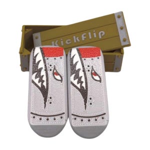 May include: Two gray and red finger skateboards with a shark-like design, displayed in front of a wooden box with the word "Kickflip" on it. The skateboards have a detailed pattern and are designed for finger skateboarding.