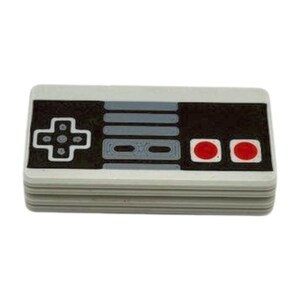 May include: A stack of white coasters featuring a retro video game controller design. The controller has a black background with gray buttons, a directional pad, and two red circular buttons. The design is a nostalgic nod to classic gaming.