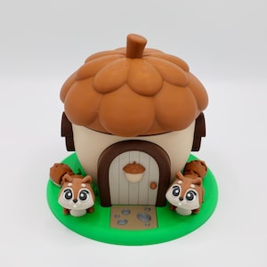 May include: A whimsical, handcrafted acorn-shaped house with a brown cap and a cream-colored base. The house features a small door and is set on a green base, flanked by two adorable squirrel figurines. A charming decorative piece.