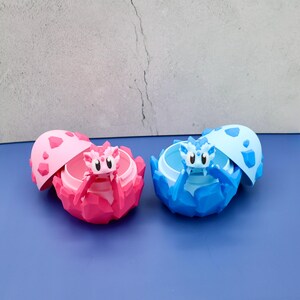 May include: Two dragon-themed trinket boxes, one pink and one blue, each with a matching lid. The boxes are shaped like cracked eggs, with a small dragon figure inside. The pink box has a pink dragon, and the blue box has a blue dragon.