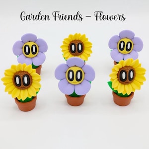 May include: Six whimsical flower figurines in terracotta pots. The flowers are either yellow sunflowers or purple daisies, each with a smiling face and the number "00" for eyes. The text "Garden Friends - Flowers" is above the figurines.