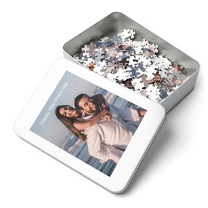 May include: A white metal puzzle tin with an open lid, revealing a pile of puzzle pieces. The tin's lid displays a photo of a couple on a beach, with the text "Happy Valentine's Day". The puzzle pieces are various shades of gray.