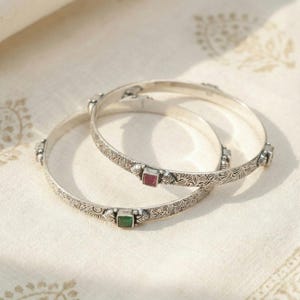Puede incluir: Dos brazaletes de plata con intrincados patrones grabados y detalles de piedras preciosas cuadradas. Un brazalete presenta una piedra preciosa roja, el otro una piedra preciosa verde. Las joyas se exhiben sobre una superficie color crema.
