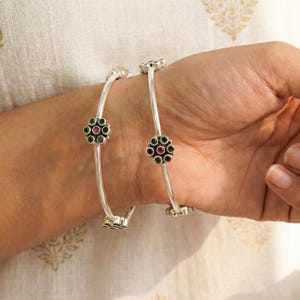 Può includere: Due braccialetti d'argento con disegni floreali. Ogni braccialetto presenta piccole pietre preziose verdi e rosse disposte a forma di fiore. I braccialetti sono mostrati su un polso, evidenziando il loro design elegante.