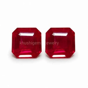 May include: Two radiant-cut, deep red gemstones. The gems are square with beveled edges, reflecting light. The image is set against a white background. The text "khushigem jewelry" is visible on the gems.