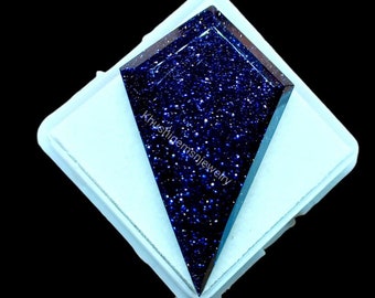 May include: A kite-shaped blue sandstone gemstone with a sparkling, glitter-like texture throughout its surface. The dark blue stone features the text Khushigemsnjewelry printed across its center and is set against a white square base.