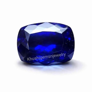 May include: A deep blue, cushion-cut sapphire gemstone. The gem is highly faceted, reflecting light, and has a high degree of clarity. The text "Khushigemsnjewelry" is visible on the bottom of the gem.