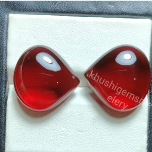 May include: Two teardrop-shaped, polished red gemstones. The gems have a glossy finish and a lighter, reflective spot near the top. The text "khushigemselelry" is visible on one gem. They are displayed in a black and white case.