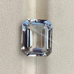 Puede incluir: Una gema rectangular transparente con talla esmeralda. La gema tiene un tono azul claro y está facetada para reflejar la luz. El texto "khushigemsewelry" es visible en la gema. La piedra preciosa está sobre un fondo neutro.