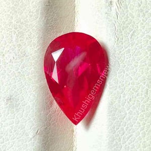 Puede incluir: Una gema de rubí facetada en forma de pera. La gema es de color rojo vibrante con múltiples facetas que reflejan la luz. La piedra preciosa está sobre un fondo texturizado blanquecino. El texto "Khushigemssnjew" es visible en el lado derecho.