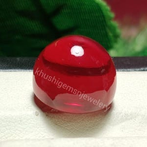 Peut inclure: Une pierre précieuse rouge translucide et brillante, de forme arrondie et taillée en cabochon. Elle présente une petite inclusion blanche. Le texte "khushigemsnjewellery" est visible. La gemme est posée sur un fond neutre.