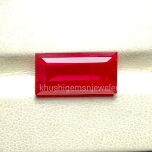 Könnte beinhalten: Ein rechteckiger, facettierter, tiefroter Edelstein mit dem weißen Text "khushigemsnjeweler". Der Edelstein ist vor einem weißen, strukturierten Hintergrund platziert, was auf eine Schmuckpräsentation hindeutet.