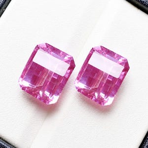 May include: Two rectangular, faceted pink gemstones with a white band across the center. The gems are set against a white background, suggesting a jewelry display.