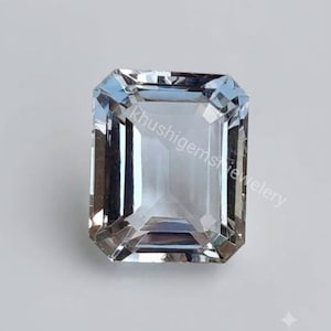 May include: A rectangular, emerald-cut, light blue gemstone with faceted edges. The stone is transparent and reflects light, featuring the text "khushigemsjewelery" overlaid diagonally across the centre of the stone's surface.