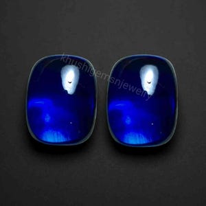 Puede incluir: Dos piedras preciosas rectangulares de color azul intenso con esquinas redondeadas y una superficie de cabujón lisa y pulida. Las piedras muestran un tono azul vibrante y saturado con reflejos de luz blanca brillante. Texto: khushigemsnjewelry.