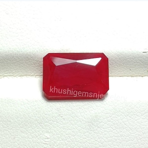 Könnte beinhalten: Ein facettierter, rechteckiger roter Edelstein mit dem Text "khushigemssn jewelry" auf der Unterseite. Der Edelstein hat eine polierte Oberfläche und ist vor einem weißen Hintergrund platziert. Der Edelstein ist ein tiefes, leuchtendes Rot.