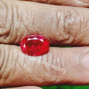 May include: A faceted, oval-shaped, bright red gemstone. The gem is held between fingers, with the text "Khushigem snjewelery" visible. The gem's facets reflect light, creating a sparkling effect.