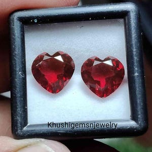 May include: Two heart-shaped, faceted, deep red gemstones are displayed in a black case with a white interior. The gemstones are highly reflective, catching the light. The text "Khushigemsnjewelry" is visible at the bottom of the case.