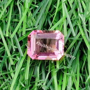 May include: A pink gemstone with a rectangular cut, set against a green grassy background. The gemstone is faceted and has a slight shimmer. The text "Khushigem" is visible in the image.