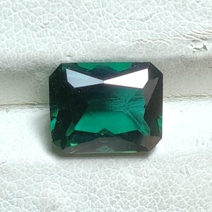 May include: A faceted, emerald-cut gemstone with a deep green hue. The gem has a rectangular shape with beveled edges, reflecting light and creating a sparkling effect. The gemstone is set against a textured, off-white surface.
