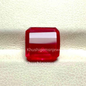 May include: A deep red gemstone with a square shape and a slightly rectangular cut. The gemstone is set on a white surface.