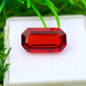May include: A vibrant, rectangular-cut gemstone in a rich red colour. The gem is displayed on a white surface, with green foliage in the background. The gemstone's cut has multiple facets, enhancing its sparkle.