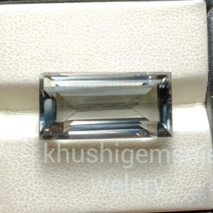 May include: A rectangular gemstone with a smoky gray hue, set against a white textured surface. The gem has a faceted cut, reflecting light. The text "khushigems" is visible on the surface. The gem is displayed in a black box.