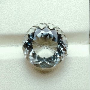 Puede incluir: Una gema clara y facetada de forma redonda. La gema tiene un corte brillante, que refleja la luz y crea un efecto brillante. El texto "khushigemsnjewellery" es visible en la gema. La gema está sobre un fondo blanco.