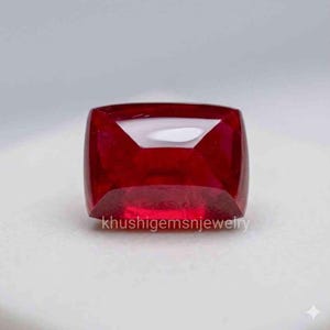 Könnte beinhalten: Ein leuchtend roter Rubin im Kissenschliff. Der Edelstein ist vor einem hellen Hintergrund platziert, wodurch seine polierten Facetten und Klarheit hervorgehoben werden. Der Text "khushigemsnjewelry" ist unten sichtbar.